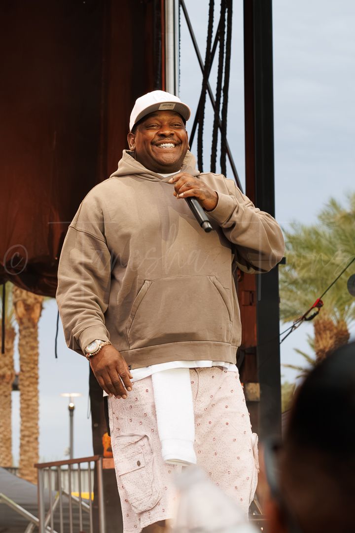 A smiling man in a beige hoodie and white cap holds a microphone on an outdoor stage with palm trees in the background.