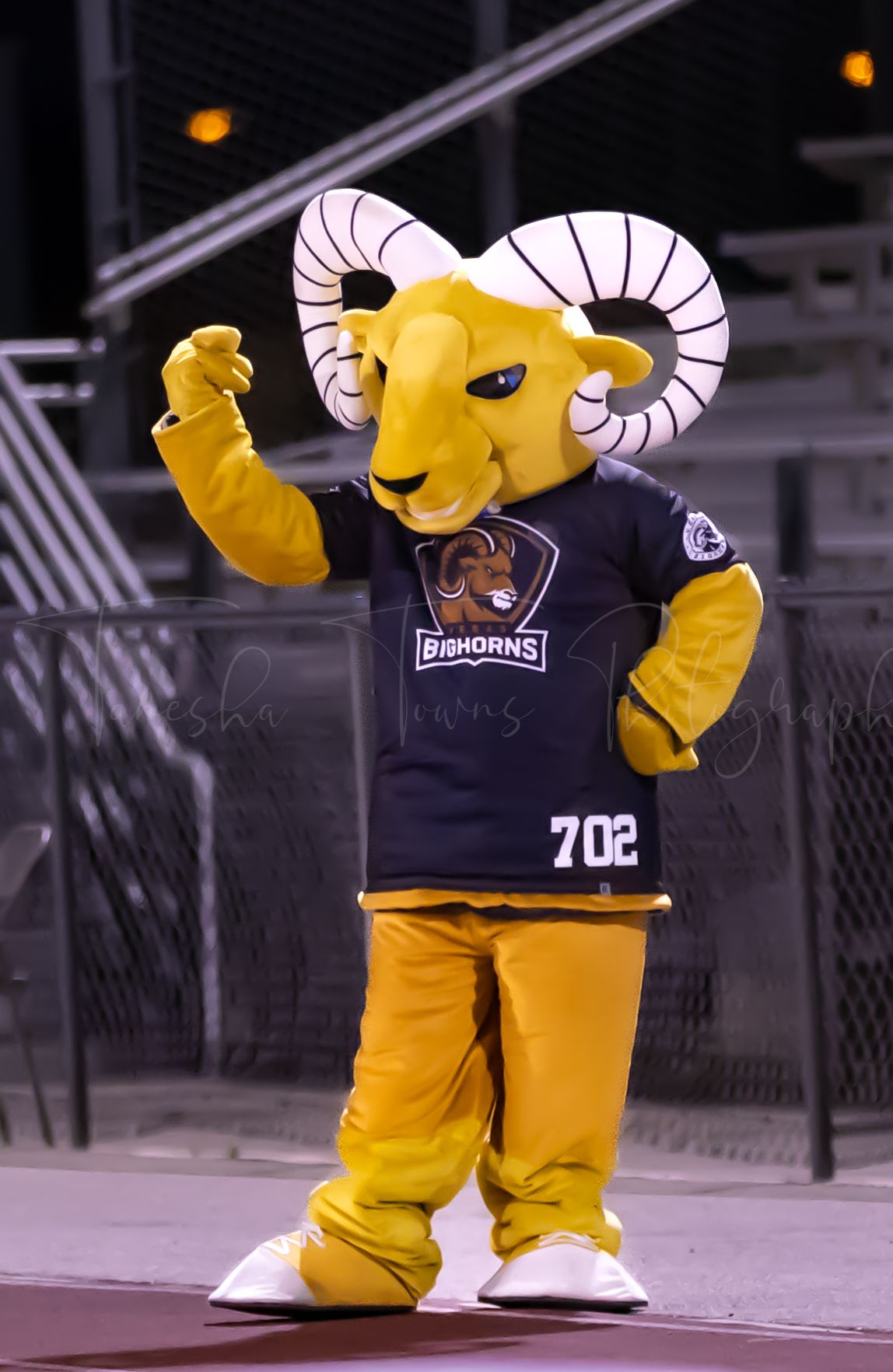Yellow ram mascot in navy uniform poses energetically at a sports event.