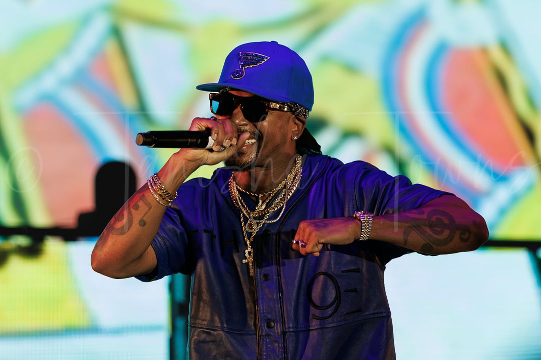 Male rapper performing on stage wearing blue cap, sunglasses, and gold chains while holding a microphone.