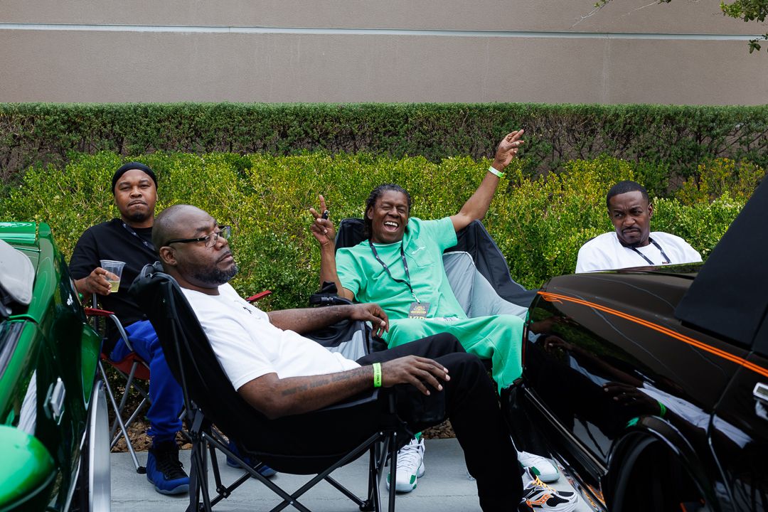 Three men relaxing in folding chairs outdoors near parked cars, one raising hands joyfully.