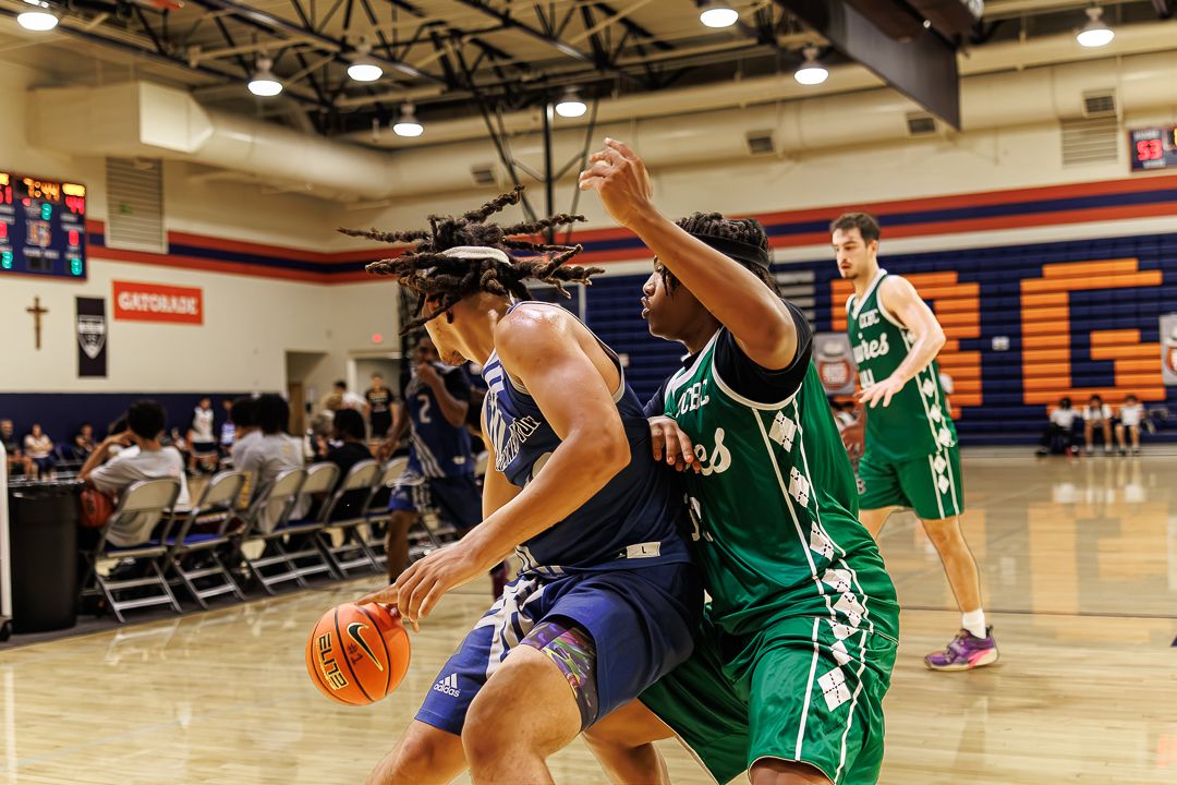 A basketball player in navy drives to the basket against a green-uniformed defender in an indoor gym.