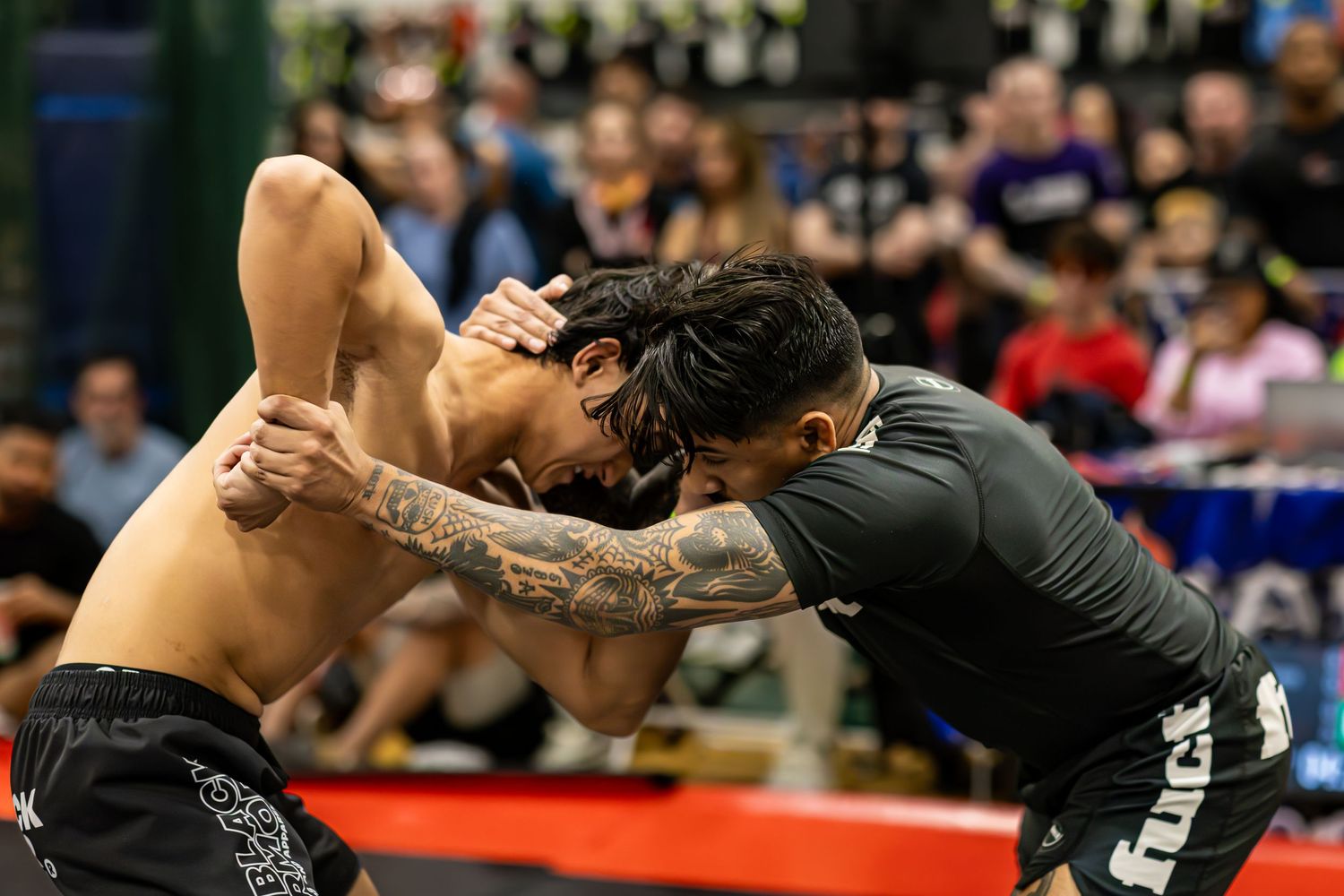 Two tattooed MMA fighters grapple intensely during a competitive submission wrestling match at an indoor tournament.