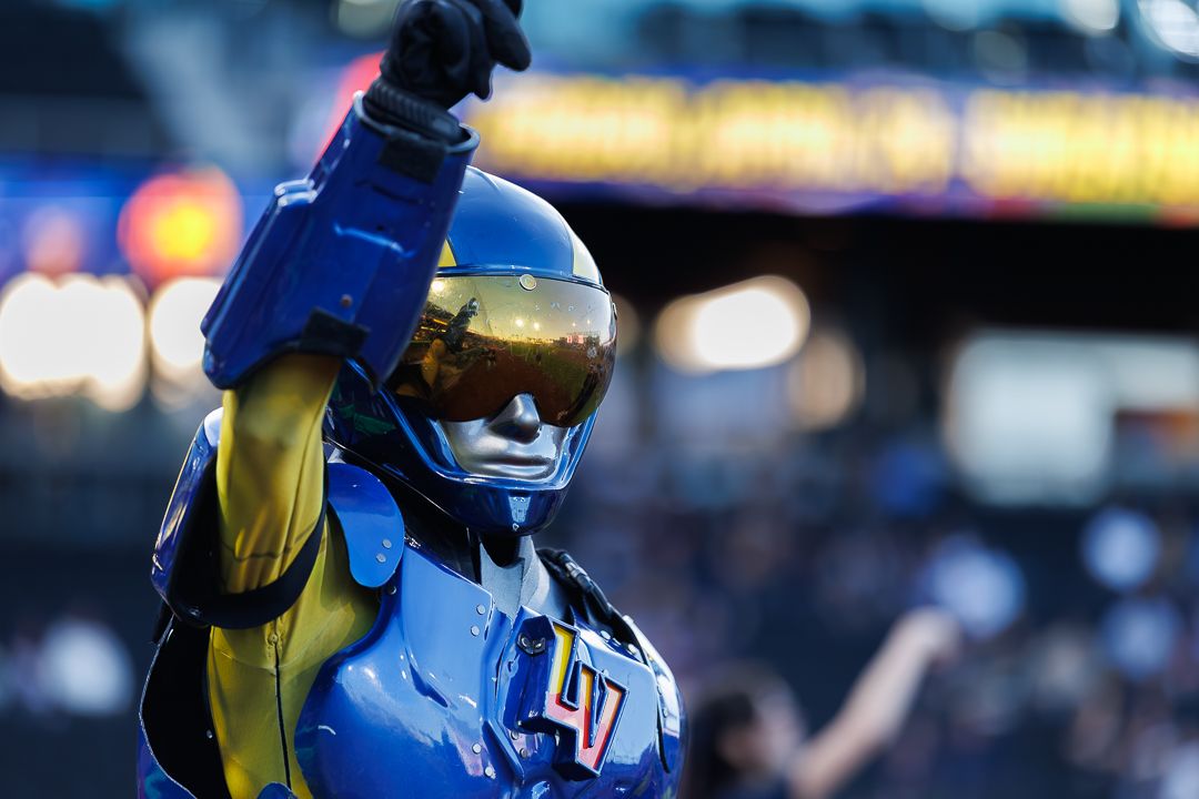 Blue and gold robot mascot in futuristic helmet raises fist at a baseball stadium event.
