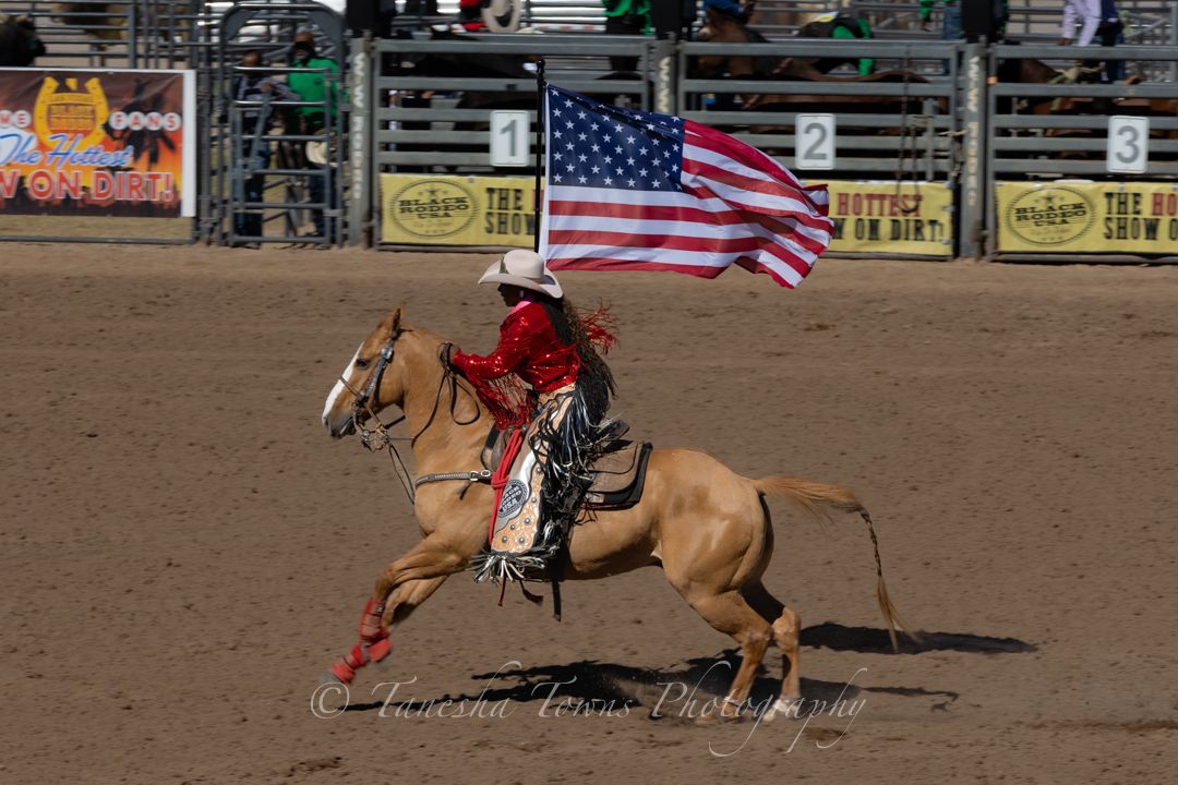 A rider carries the American flag while galloping on horseback across a dusty rodeo arena.