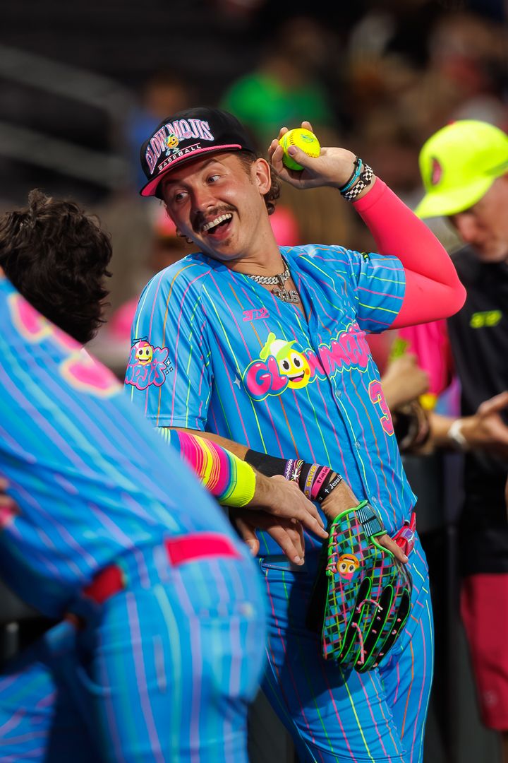 Player in colorful blue pink striped uniform holds softball aloft while smiling during a festive charity softball game.