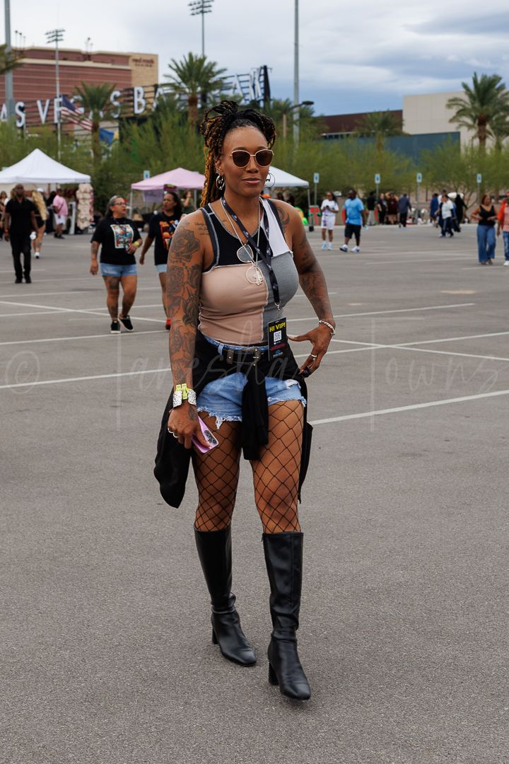 Tattooed woman in denim shorts, fishnet tights, knee-high boots, and crop top at an outdoor Las Vegas event.