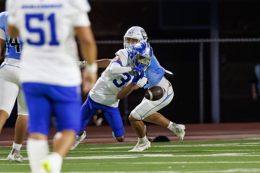 Two football players compete for the ball during a night game under stadium lights on a blue turf field.