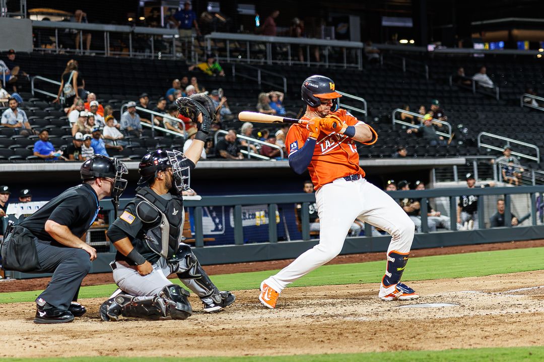 Baseball batter in orange uniform swings at a pitch as catcher and umpire watch during a night game.