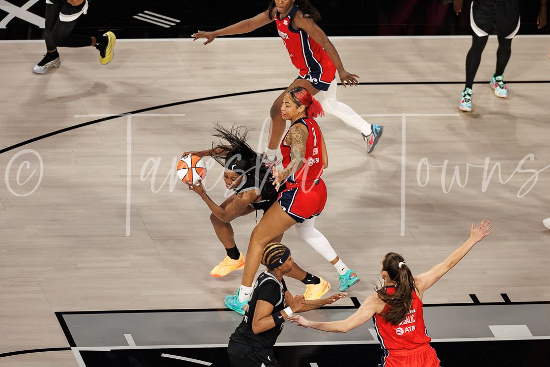 WNBA players battle for the ball during an intense basketball game, with players in black and red uniforms competing.