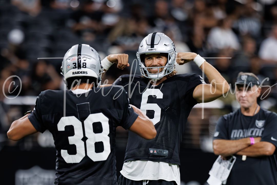 Two Raiders NFL players in black jerseys flexing on field during warm-ups at Allegiant Stadium.