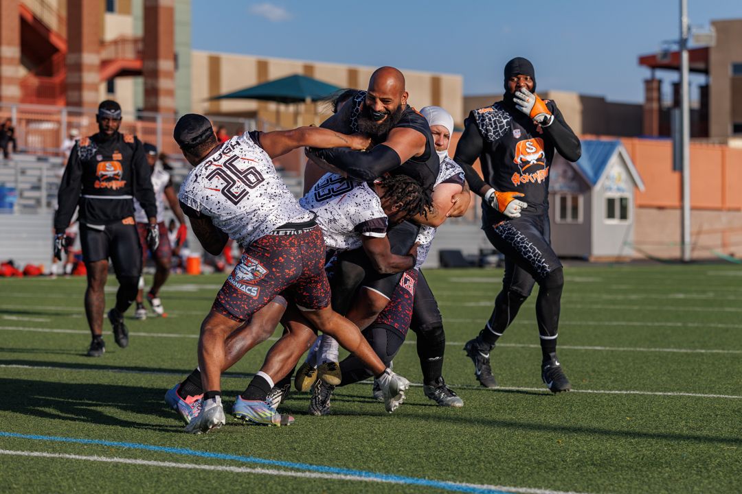 Football players collide during an intense tackle in an outdoor stadium game under clear blue skies.