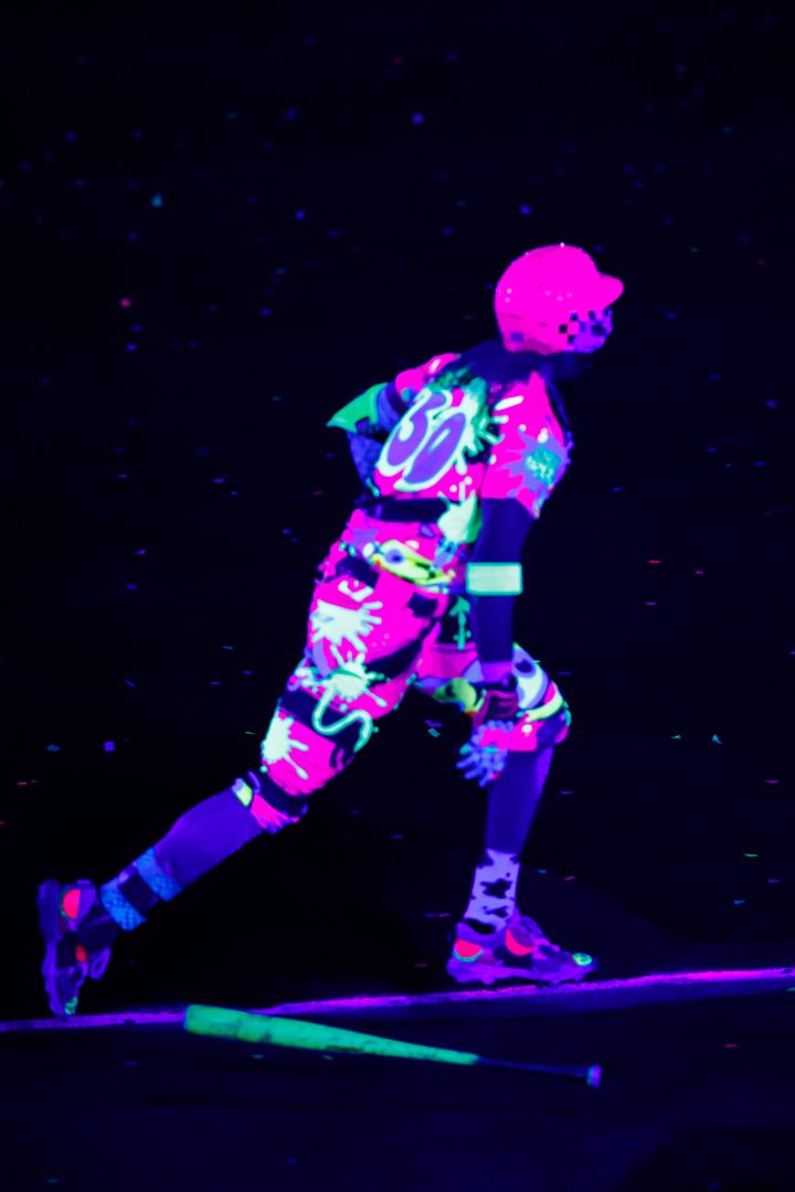 Performer in neon UV-reactive colorful outfit and pink helmet roller skating under black light.