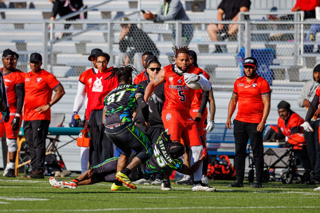 Football player in red uniform evades tackle during competitive game on stadium field with spectators watching.
