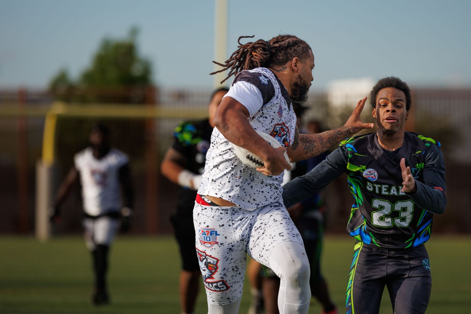Two flag football players compete on a green field, one in white and one in a green Steaz jersey number 23.