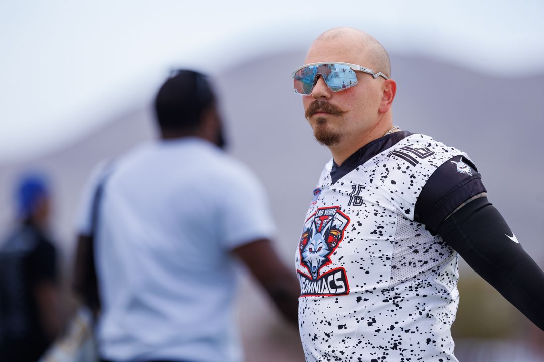Bald man with goatee wearing sunglasses and sports jersey stands on football field during outdoor practice.