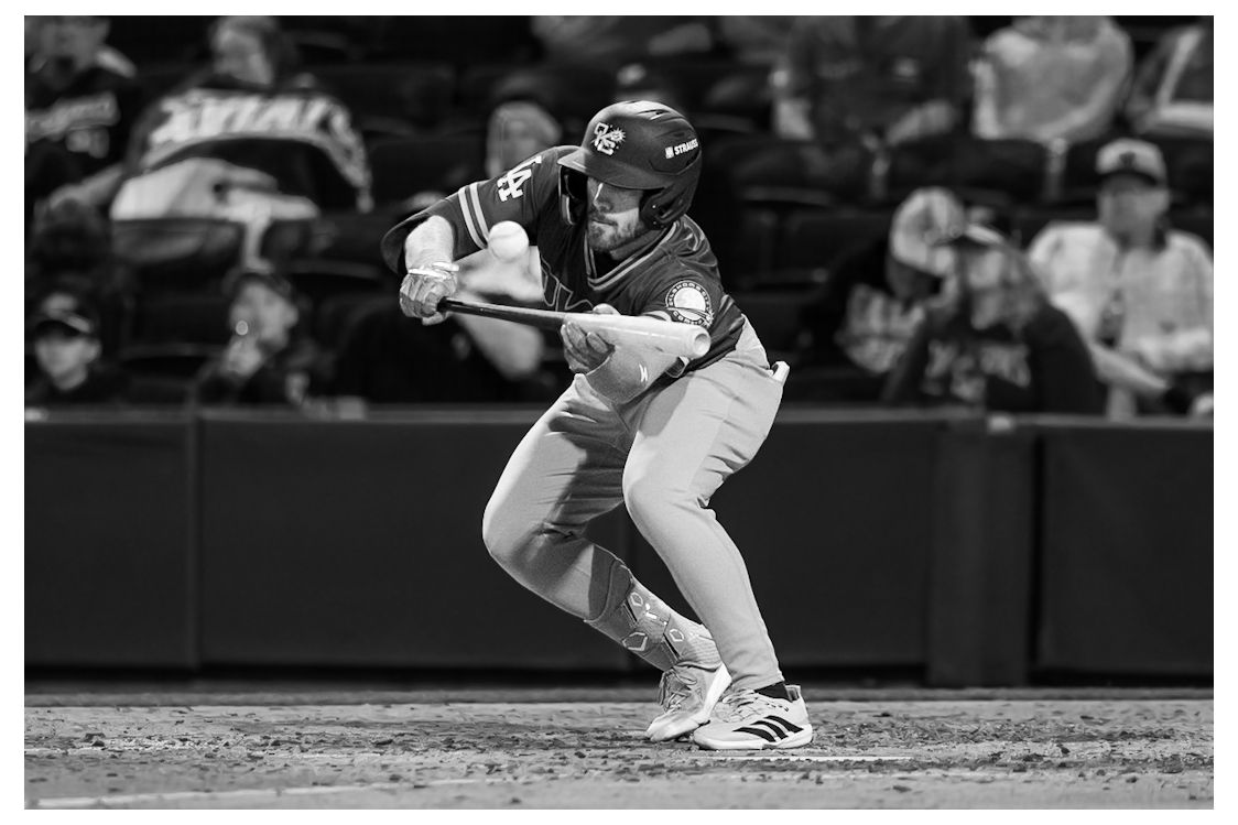 Baseball player in uniform swings bat during a night game, captured in black and white photography.
