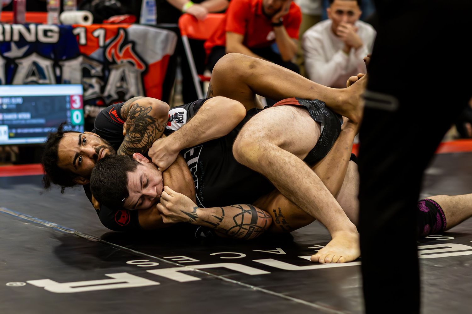 Two athletes competing in a grappling match on a black mat at an indoor wrestling tournament.