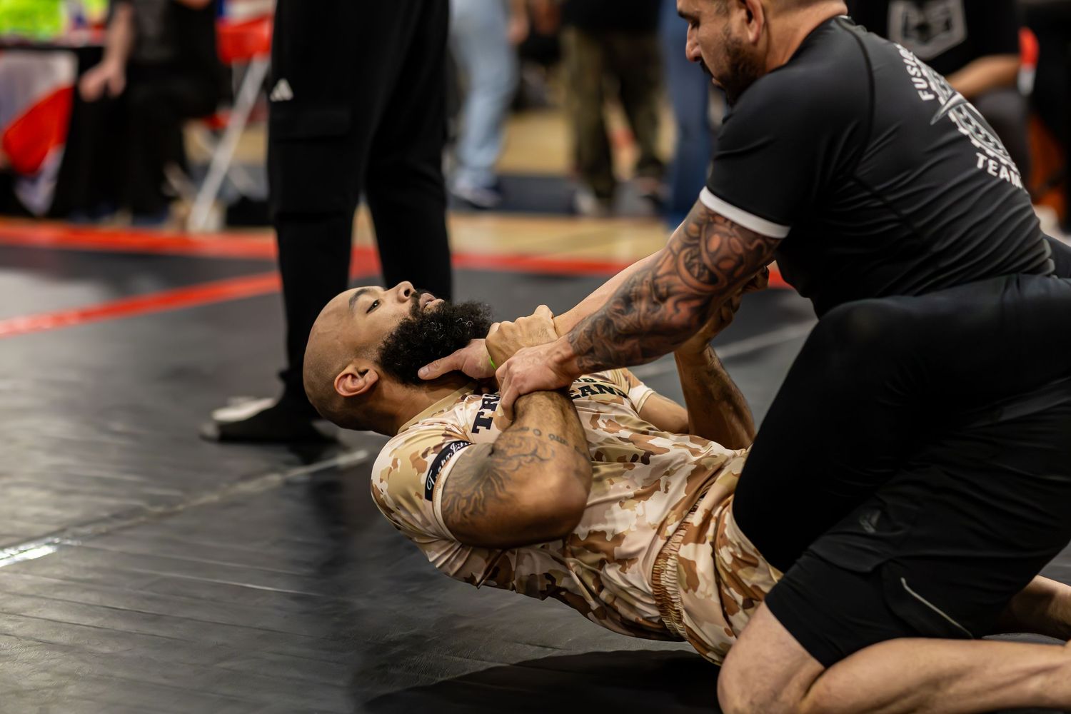 Two men competing in a grappling martial arts match on a mat at an indoor tournament.