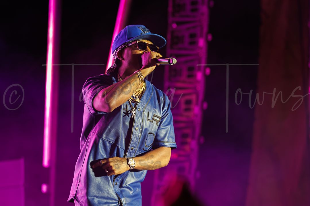 Male rapper performing on stage under purple lights, wearing a blue shirt and gold chains, holding a microphone.