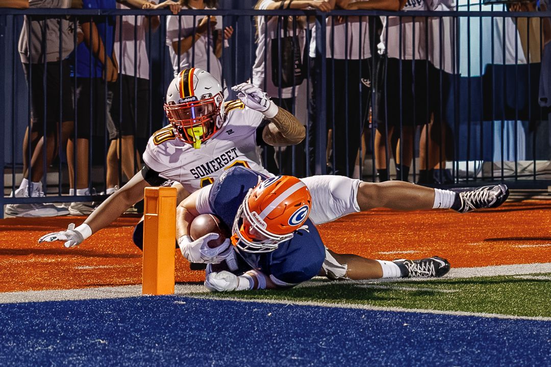 Football player dives into end zone scoring touchdown while defender attempts tackle during night game.