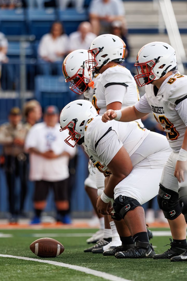 College football offensive linemen in white helmets line up at scrimmage with quarterback pointing out defensive assignments.