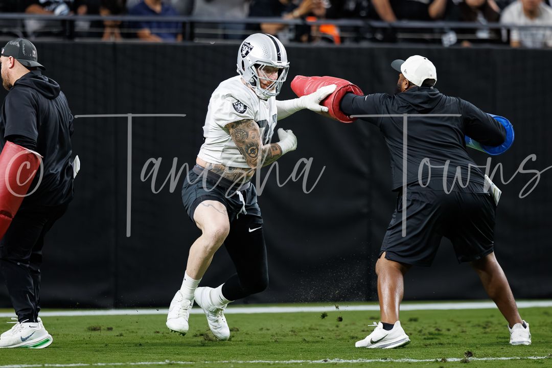 Football player in white Raiders uniform runs a drill against a coach holding a blocking pad on an indoor field.