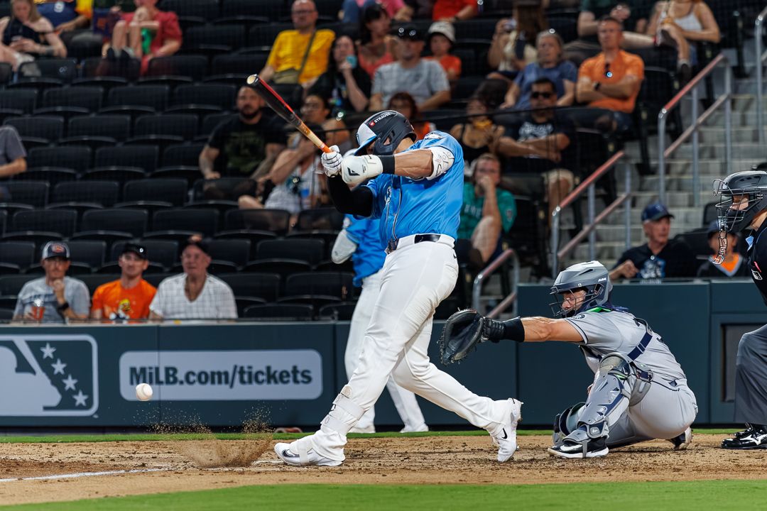 MiLB batter in blue uniform swings at pitch as catcher crouches behind home plate during night game.