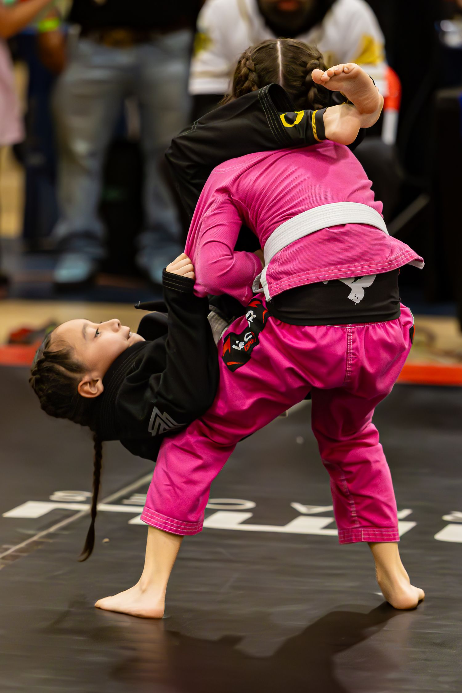 Two young girls in gi uniforms compete in a Brazilian jiu-jitsu tournament, one executing a takedown in pink gi.