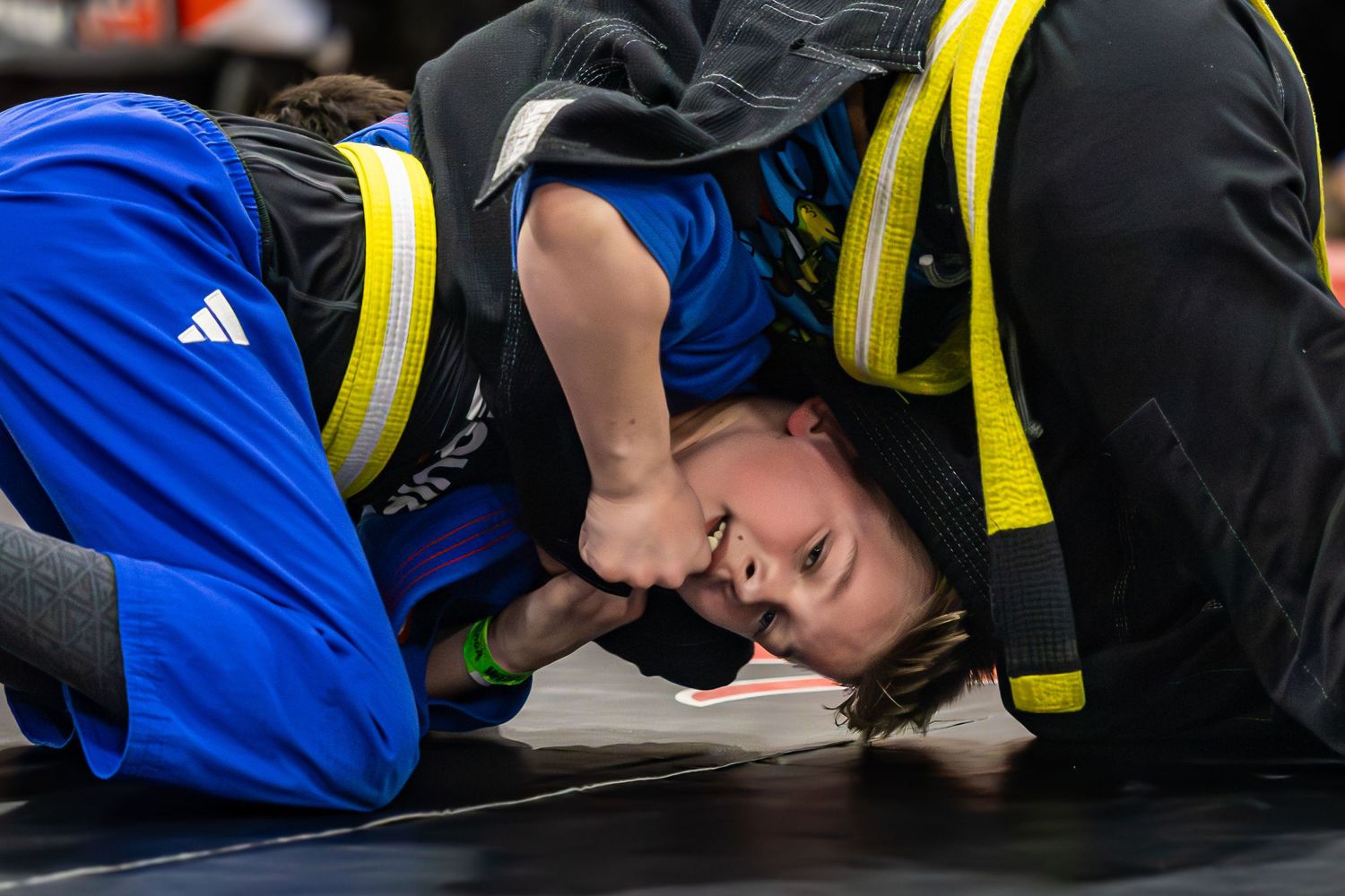 Two youth wrestlers compete on the mat, with one athlete in a blue Adidas uniform pinning their opponent down.