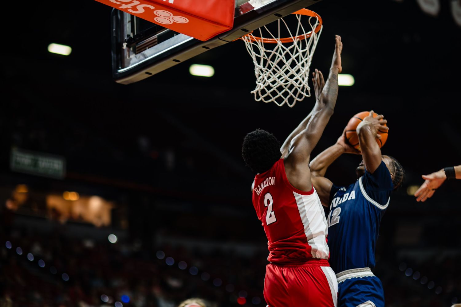 Two basketball players compete near the hoop, with one in red attempting to block a shot during an intense game.