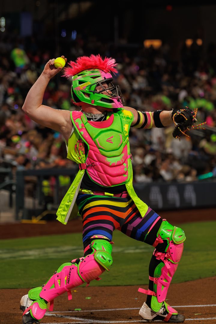 Baseball catcher in vibrant neon rainbow uniform with pink helmet throws ball during colorful themed game night.