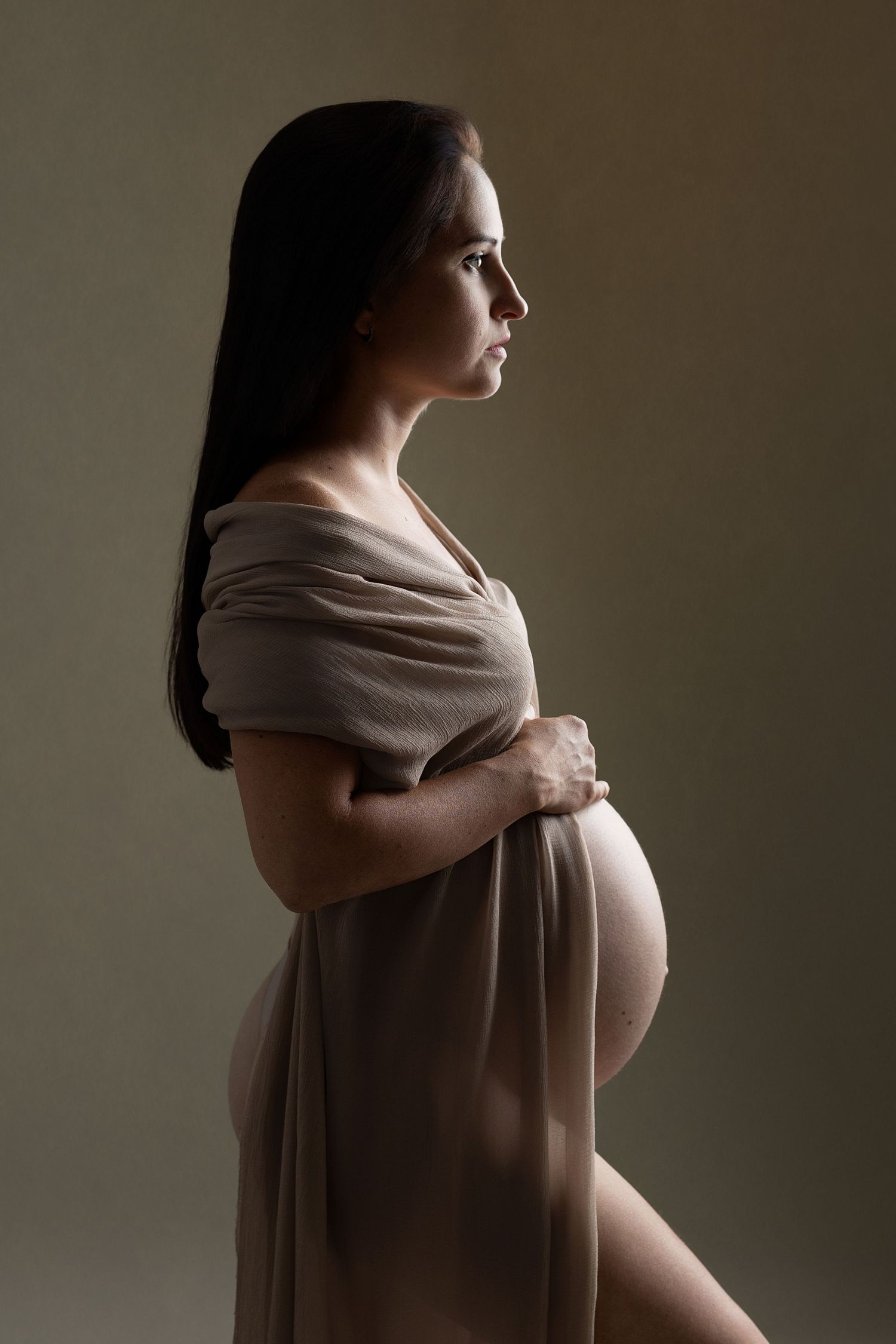Dramatic low-key maternity portraits with beige wrap against dark background.