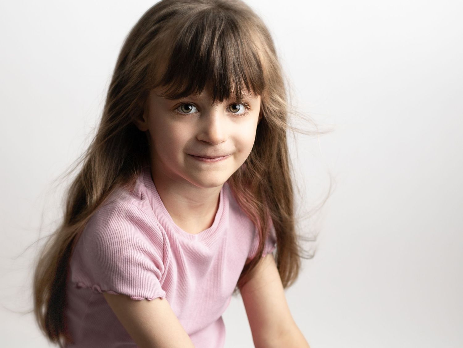 A young girl with long brown hair wearing a pink t-shirt smiles softly against a white background.