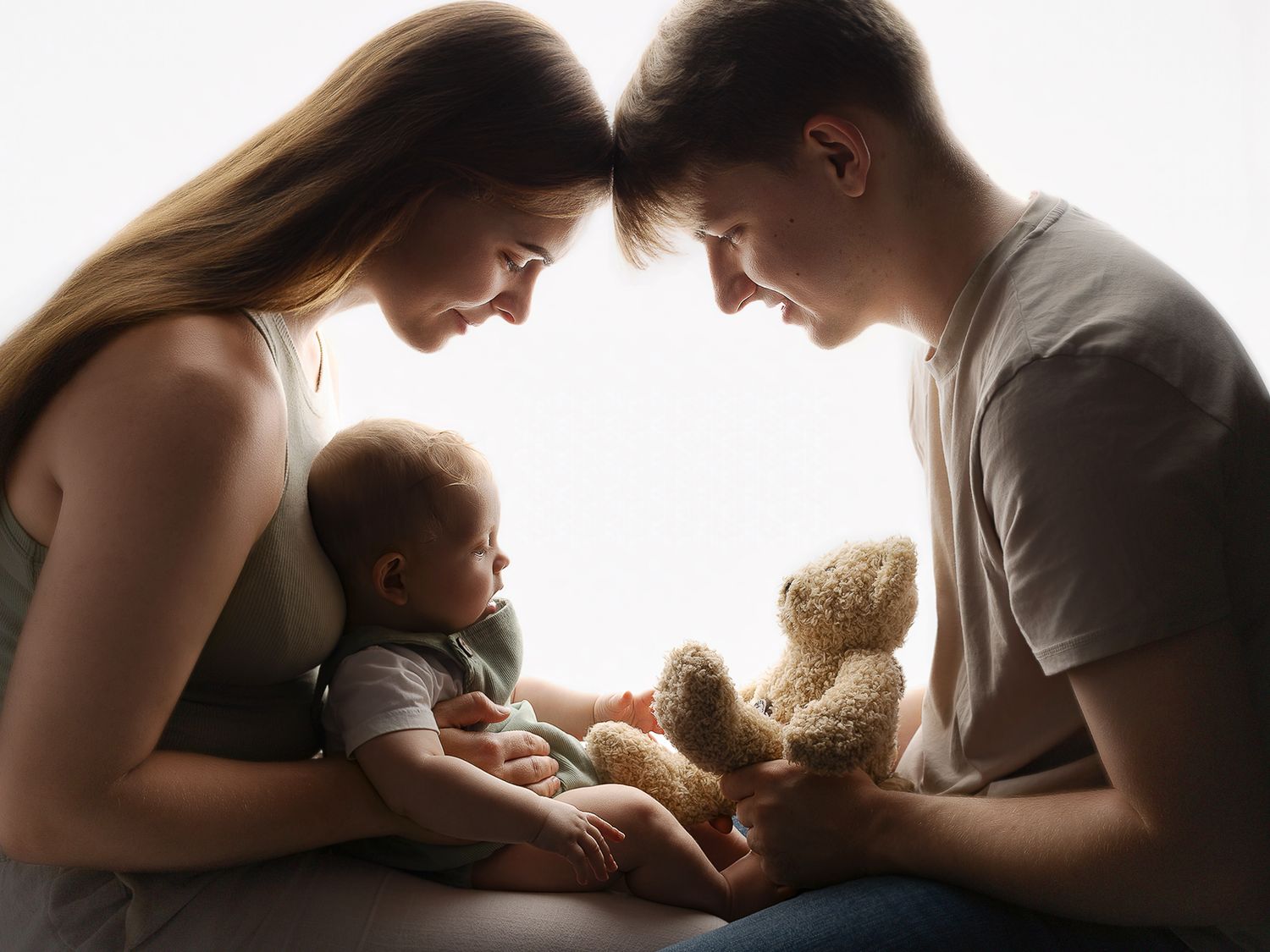 Young parents lovingly cradle their newborn baby with a teddy bear in a tender family portrait.