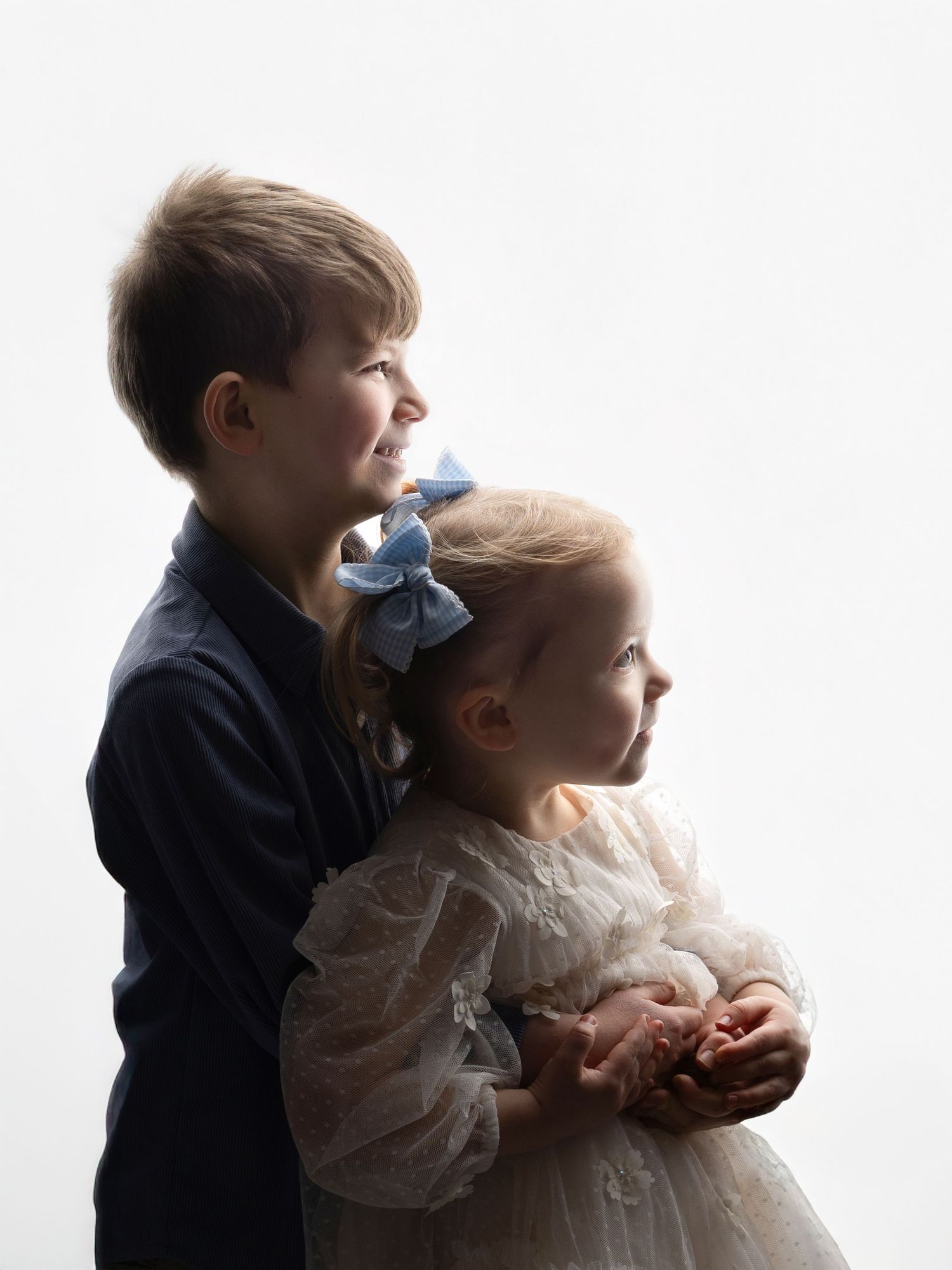 Two children in formal attire stand in profile against a white background, creating an elegant silhouette portrait.