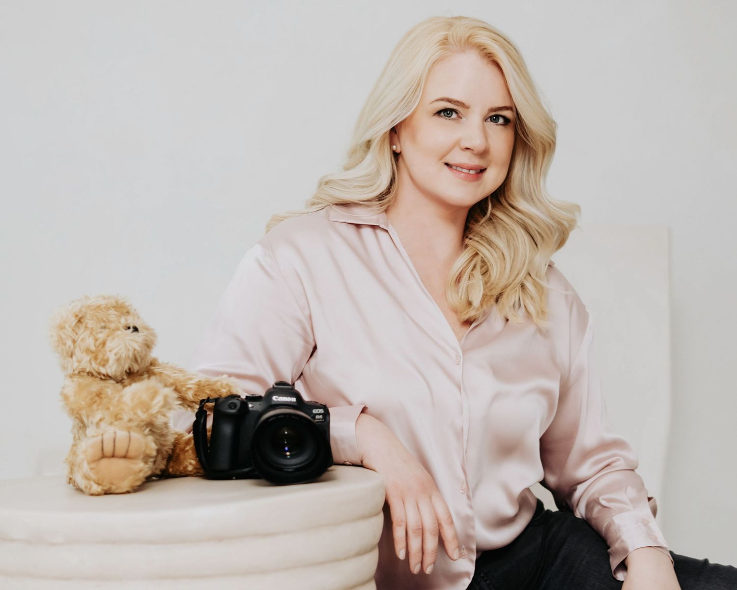 A person in a pink sweater poses with a teddy bear and camera in a light minimalist studio setting.