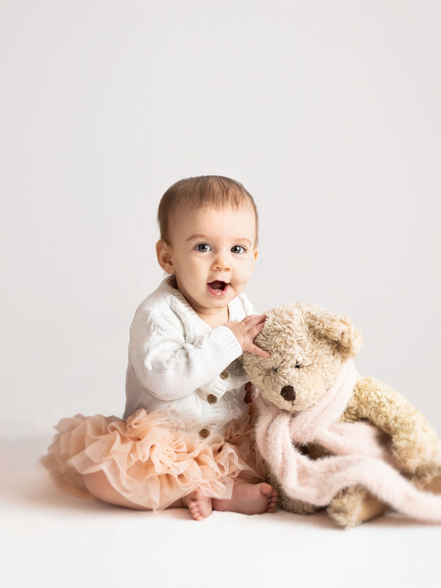 Adorable baby girl in white onesie and peach tutu sits with a teddy bear and pink blanket on white background.