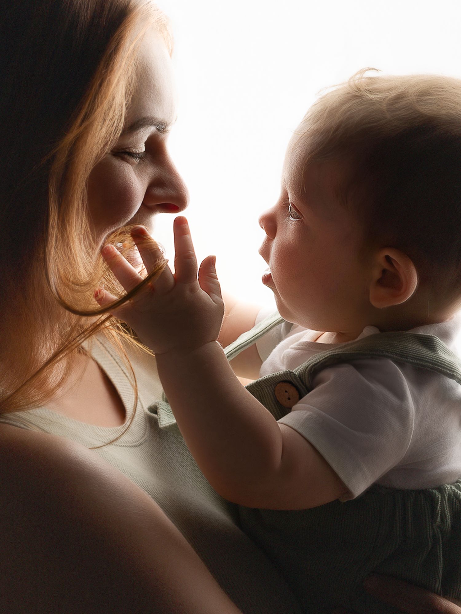 A tender moment between a smiling mother and her curious baby touching her face in soft backlit light.
