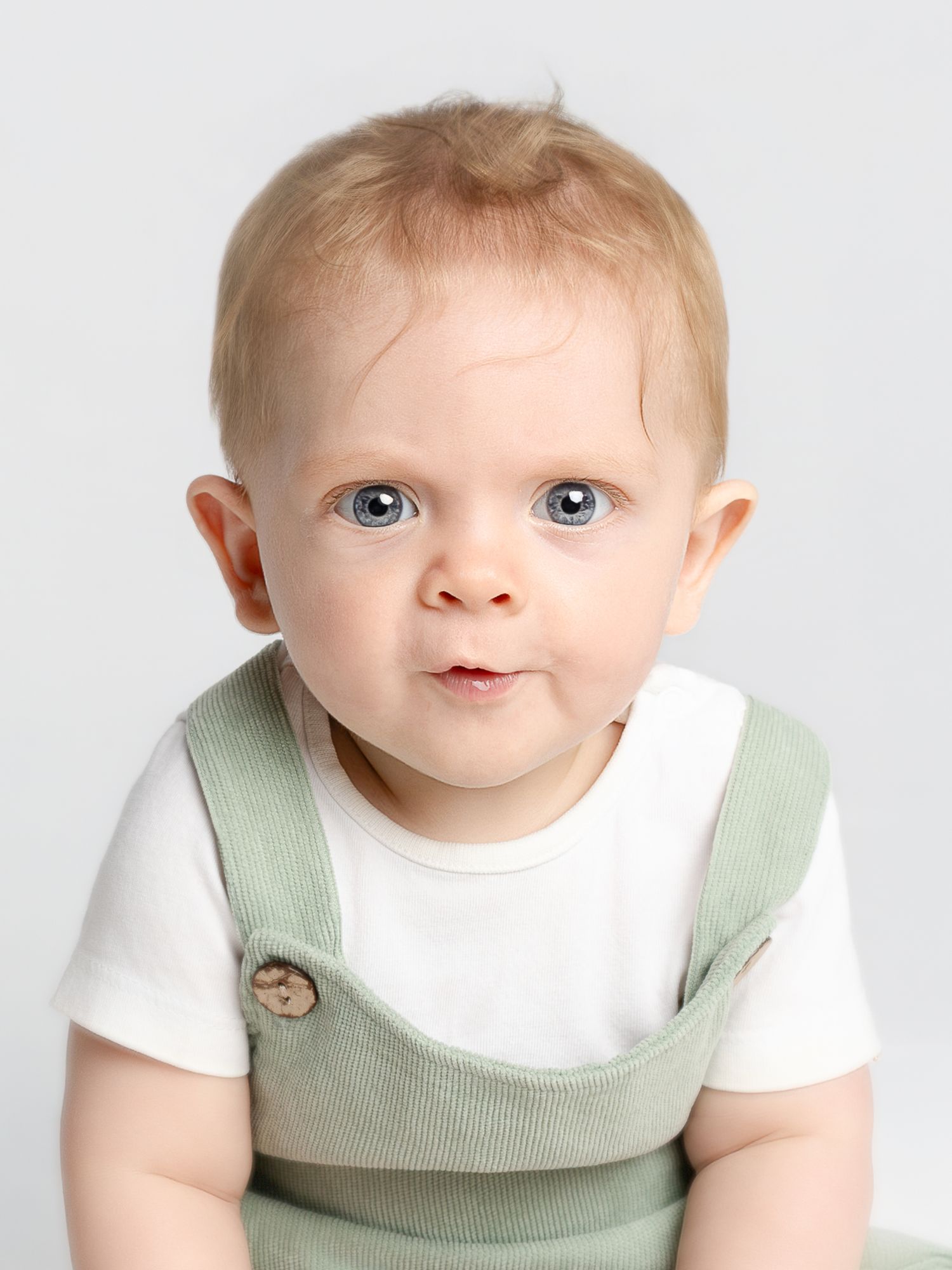 Adorable baby with blue eyes and blonde hair wearing sage green overalls and white shirt on gray background.
