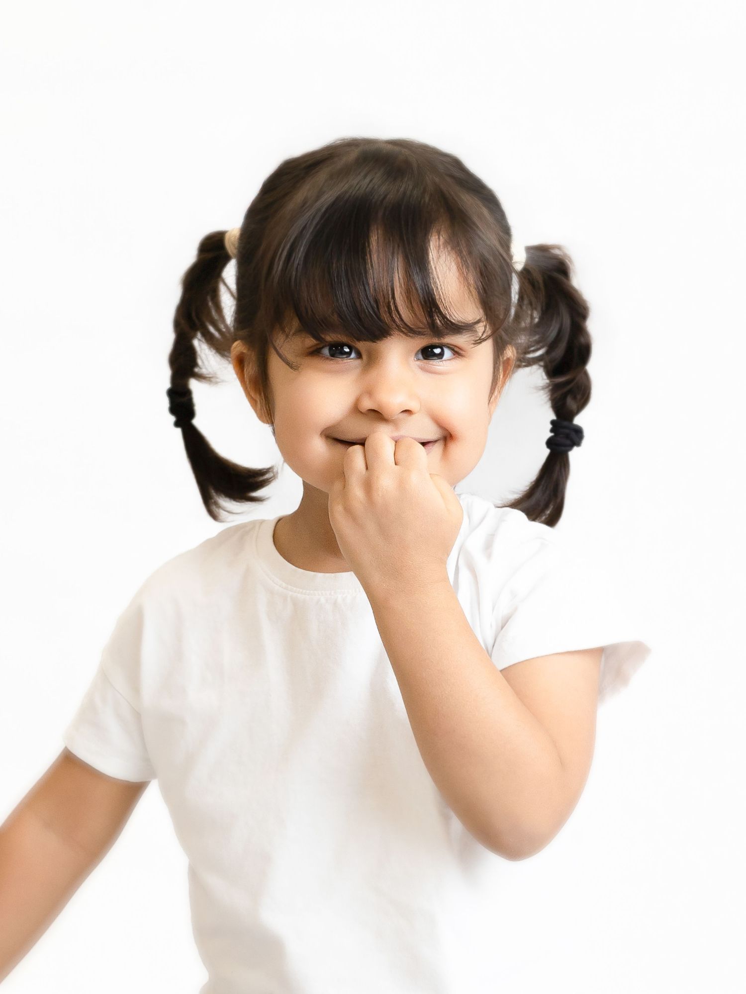 Cute little girl with pigtails in a white t-shirt covering her mouth with her hand, smiling shyly.
