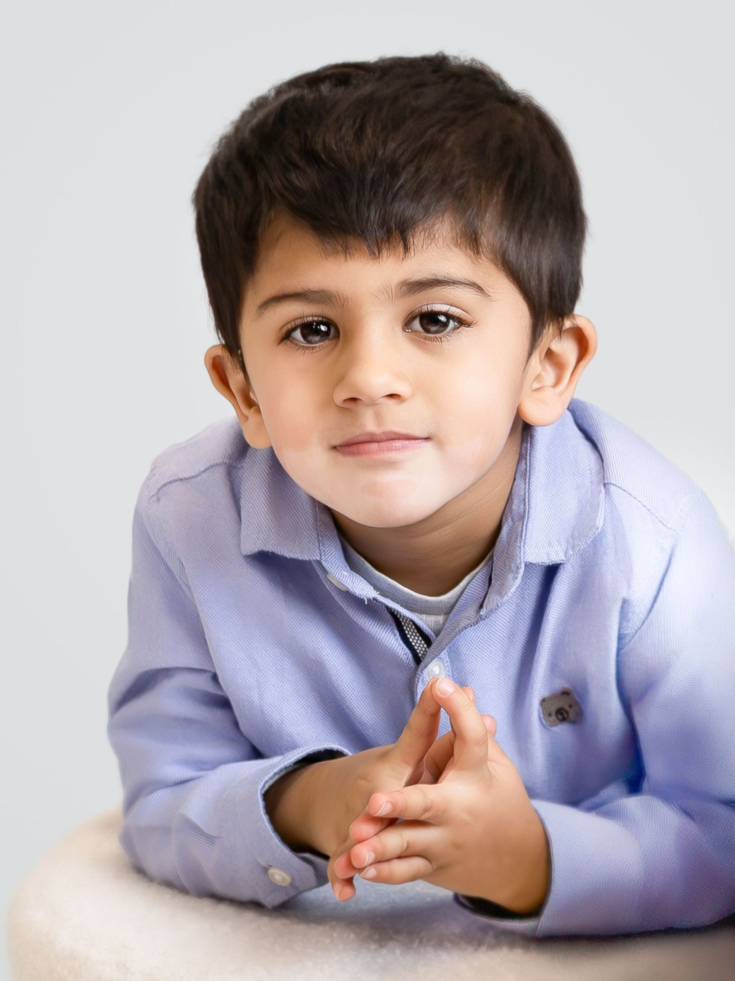 Young boy in blue shirt poses with clasped hands, looking at camera against light gray background.