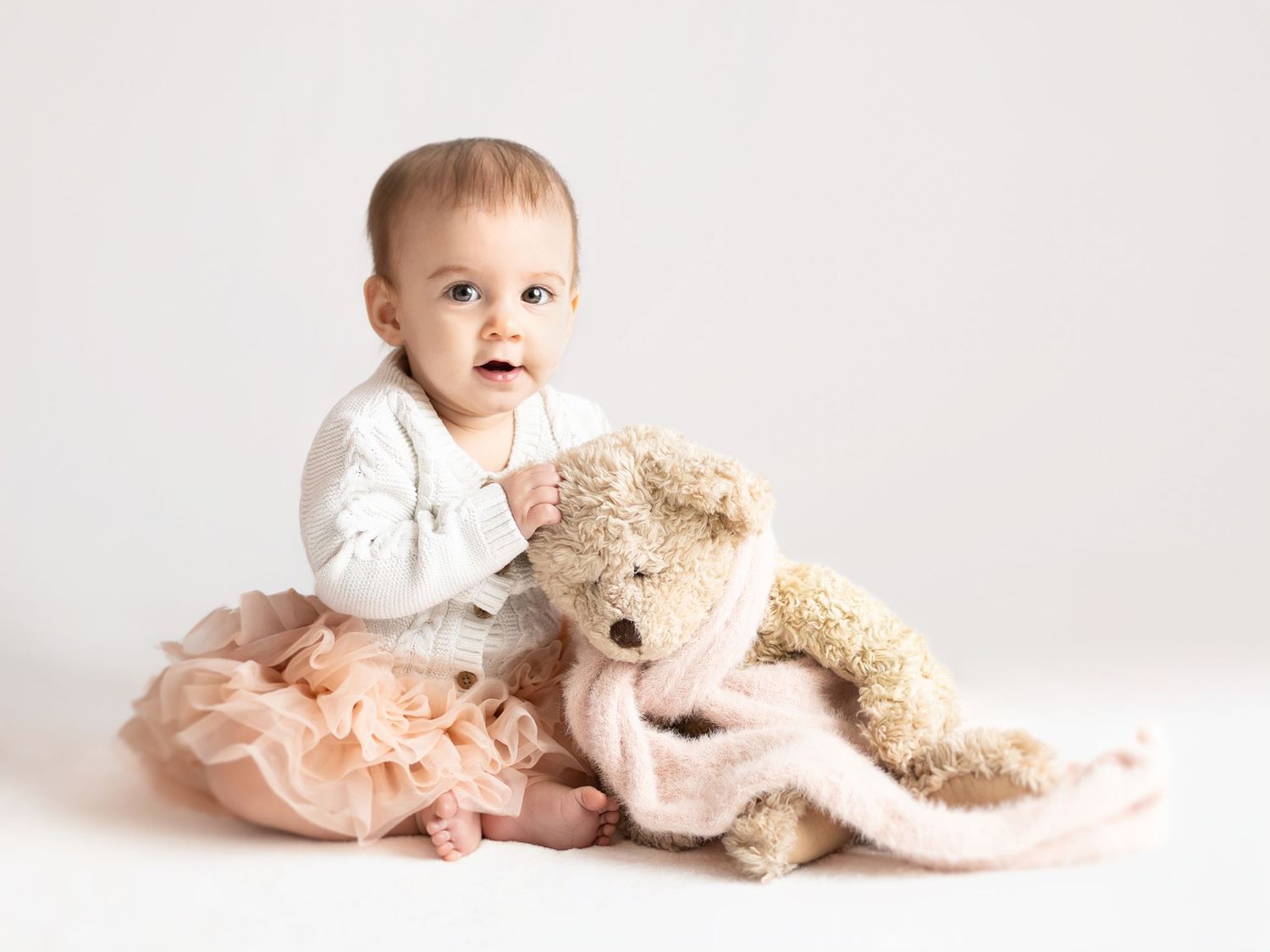 Adorable baby girl in pink tutu sits with a soft teddy bear on white background.