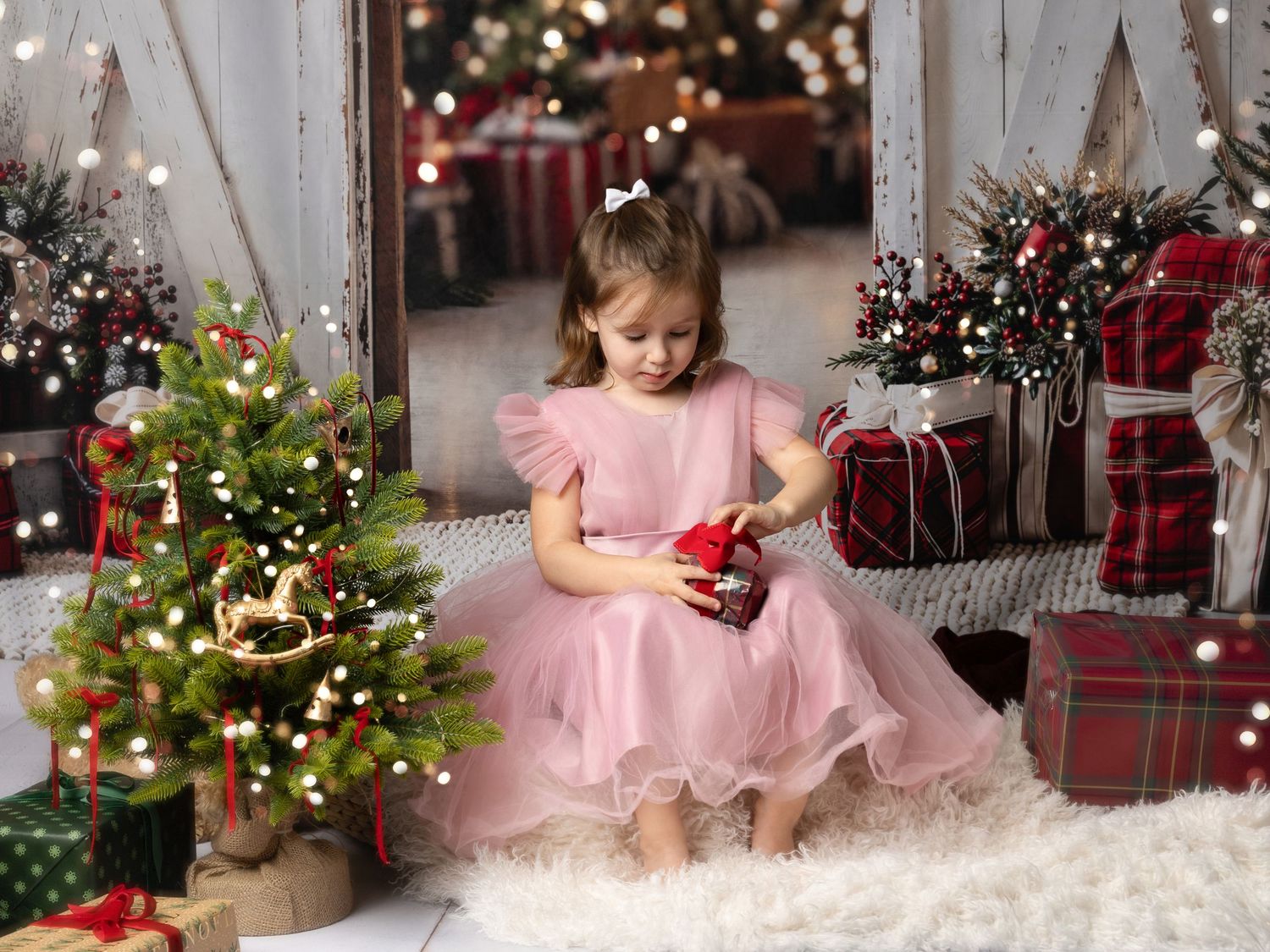 Young girl in pink dress opening Christmas gift surrounded by festive decorations, trees, and wrapped presents.
