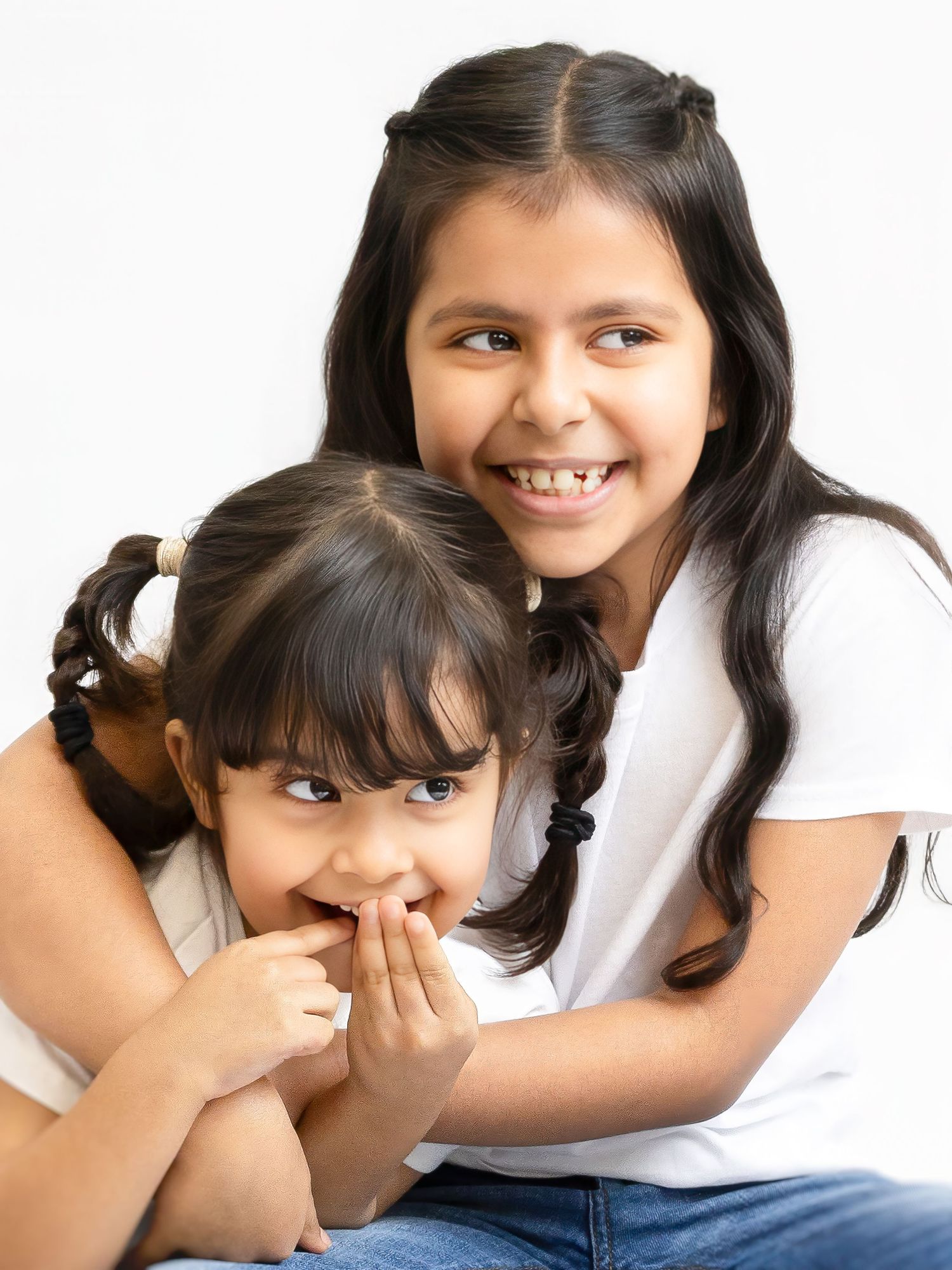 Two young sisters smiling and hugging, older girl in white tee embracing younger child, studio portrait on white background.