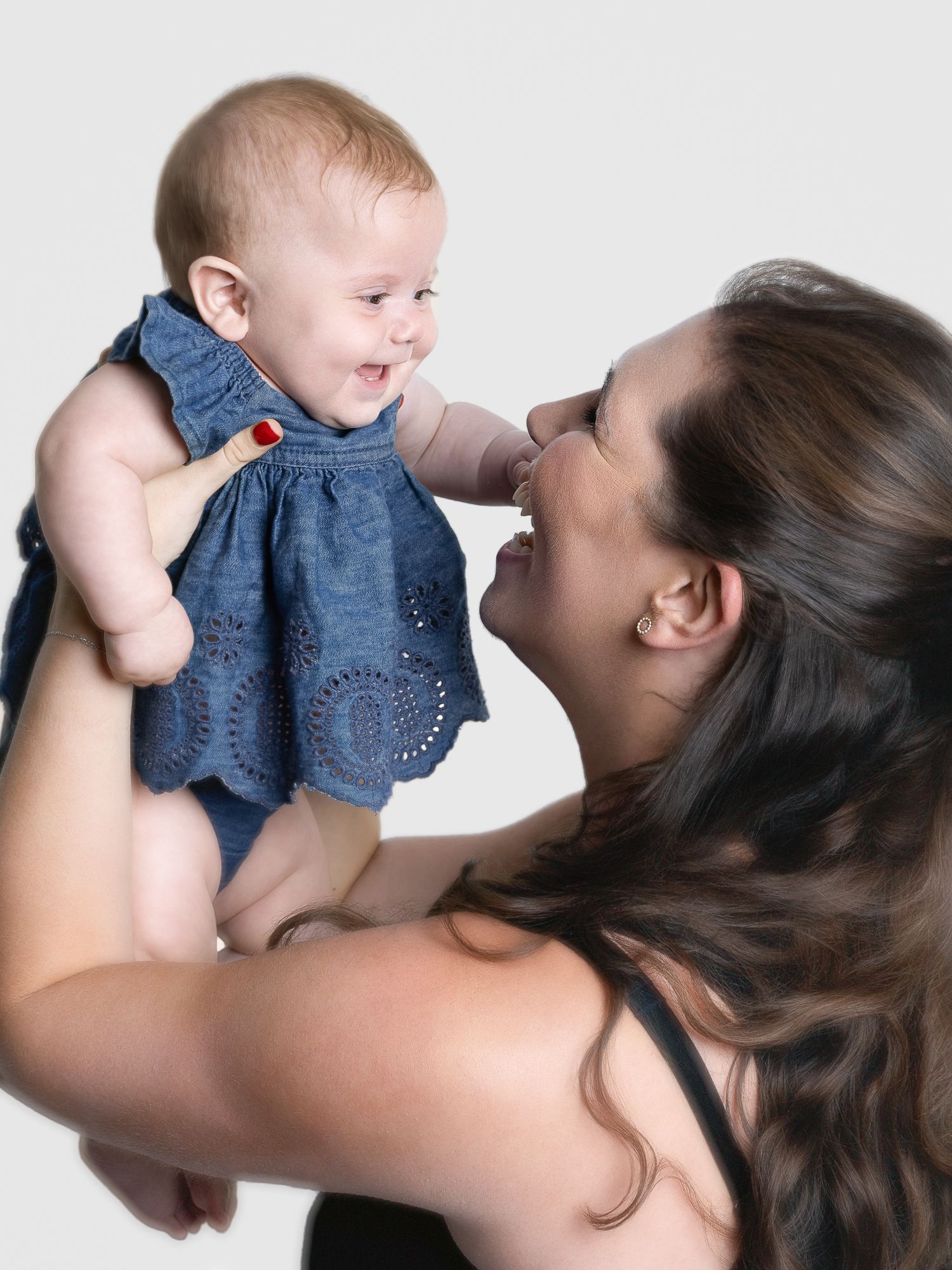 A smiling mother lifts her happy baby girl in a denim dress, sharing a joyful moment together.