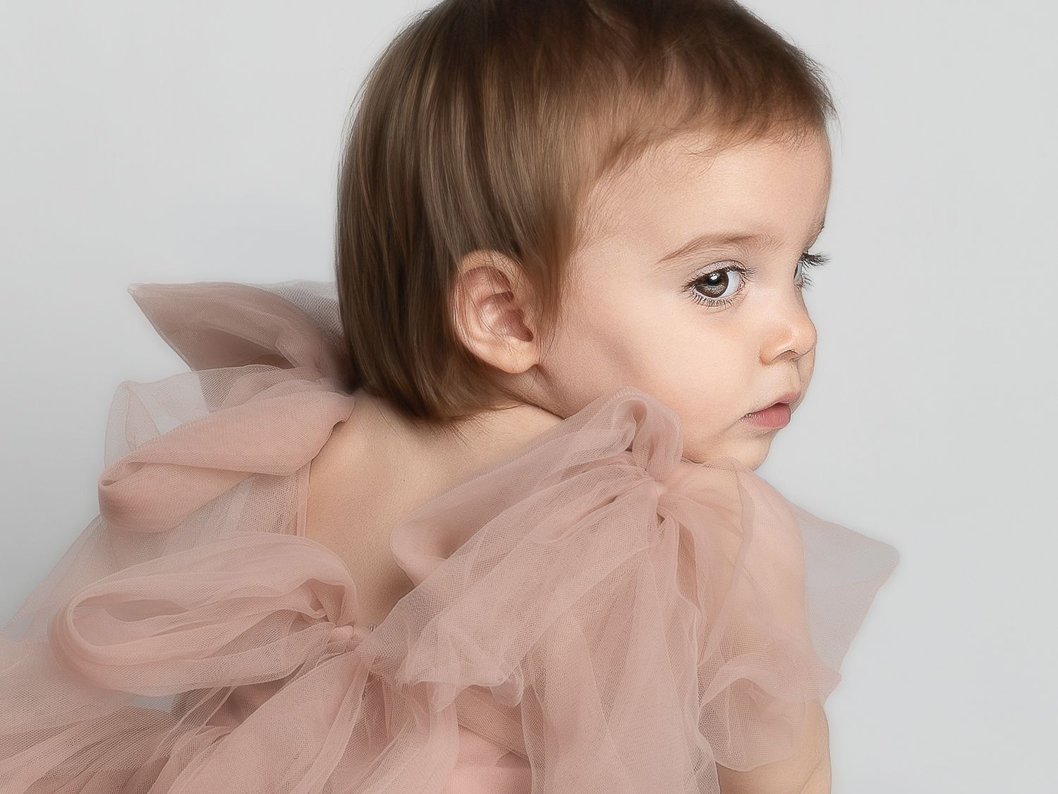 A young toddler girl in a soft pink tulle dress gazes sideways against a light gray background.