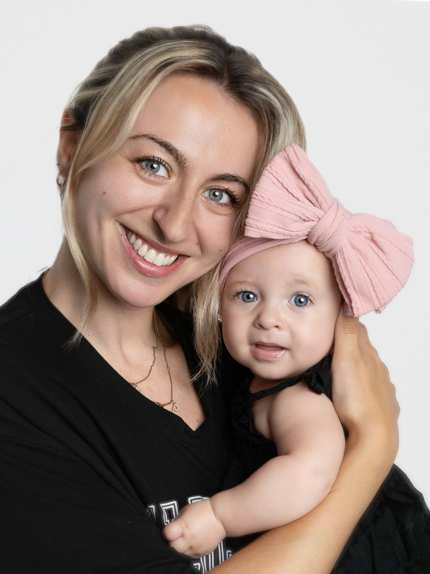 Smiling blonde woman holding a baby girl wearing a pink bow headband, both with blue eyes.