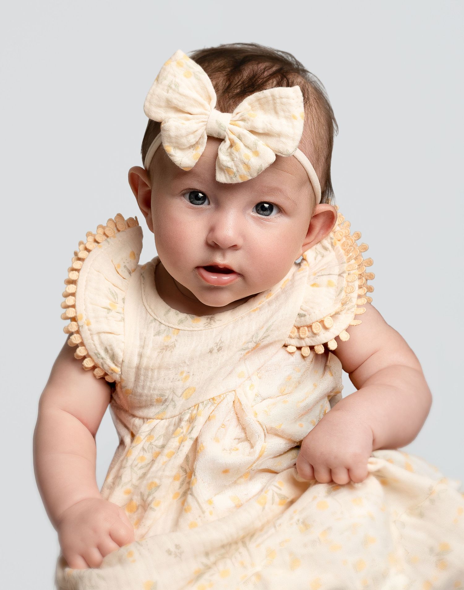Adorable baby girl in cream floral dress with ruffled sleeves and matching bow headband, sitting upright on gray background.