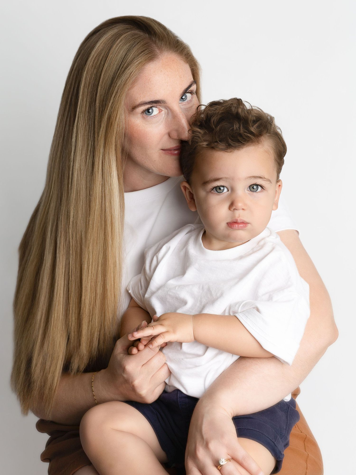 A woman with long blonde hair holds a toddler boy, both wearing white shirts, against a white background.