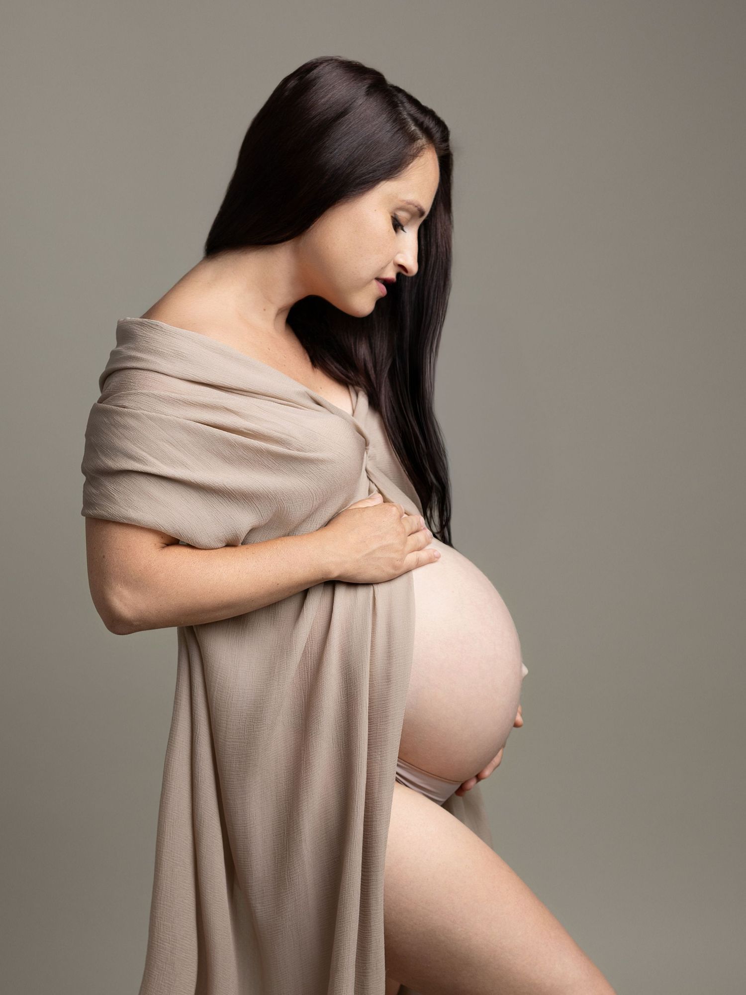 Pregnant woman in beige draped fabric holding her bare baby bump in a professional studio maternity portrait.